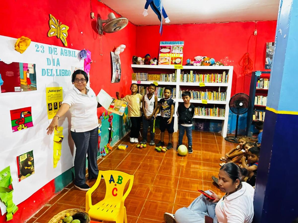 Book1 - Neighbors to Nicaragua Teacher leads a storytelling activity in a bright red library with posters and bookshelves nearby, children gathered around.