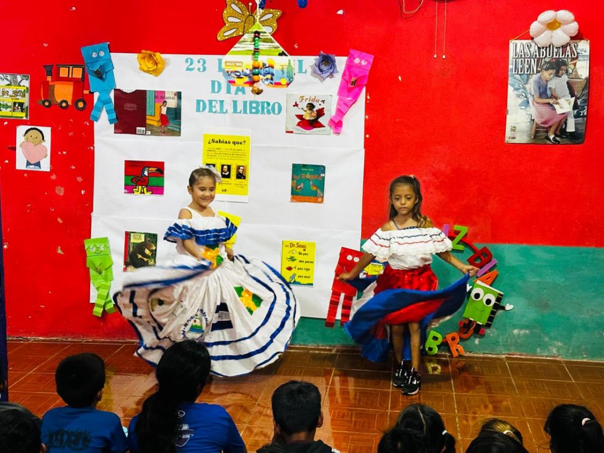 Book2 - Neighbors to Nicaragua Two young girls in colorful flamenco-style dresses dance in front of a red classroom wall; audience watches moment.