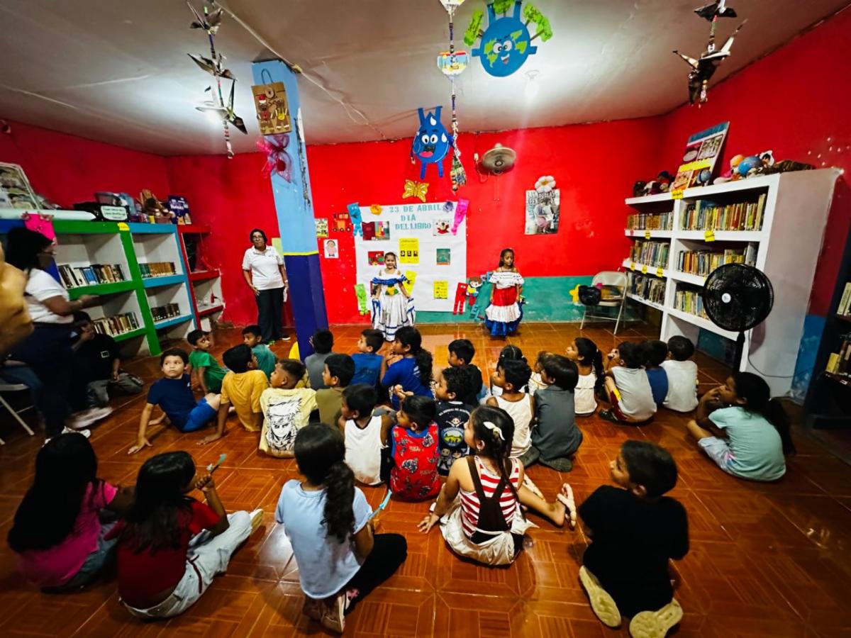 Book3 - Neighbors to Nicaragua Group of children seated on a tiled floor inside a colorful library watching two girls in traditional dresses perform at the front.