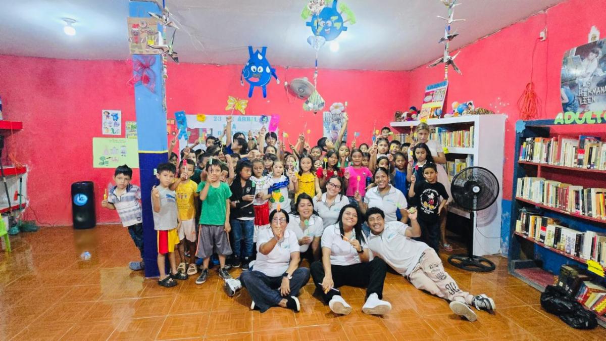 Book4 - Neighbors to Nicaragua Group of children and adults posing for a photo in a bright, decorated library/classroom with pink walls, bookshelves, and hanging crafts.