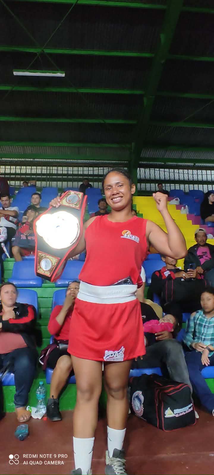 Boxing 3 - Neighbors to Nicaragua Female boxer in a red outfit holding a championship belt and posing proudly in a gymnasium with spectators in the background