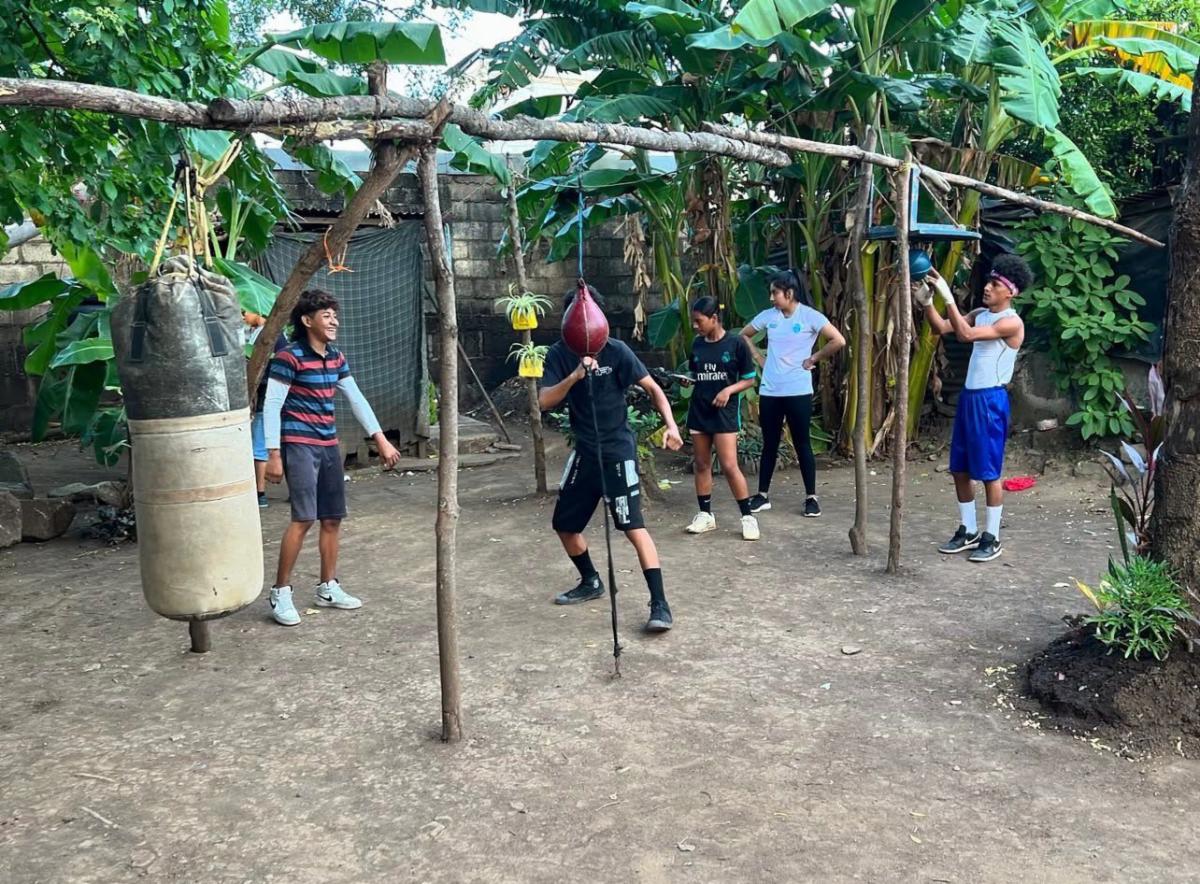 Boxing 6 - Neighbors to Nicaragua Group of youths practice boxing in a backyard, hitting a hanging punching bag under a wooden frame while others watch.
