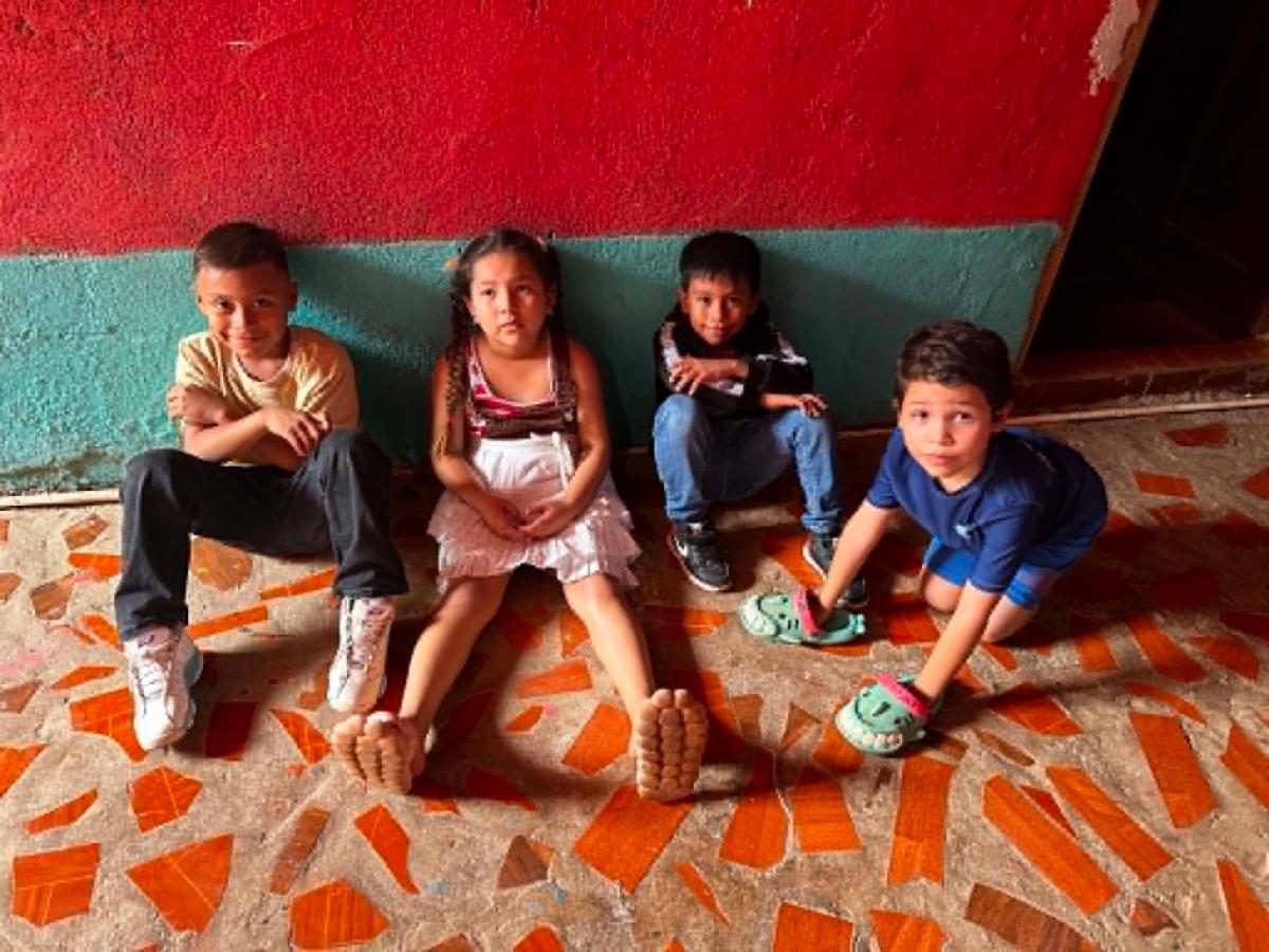 Candid1 - Neighbors to Nicaragua Four children sit against a red and teal wall on a mosaic tile floor, with bare feet extended toward the camera.