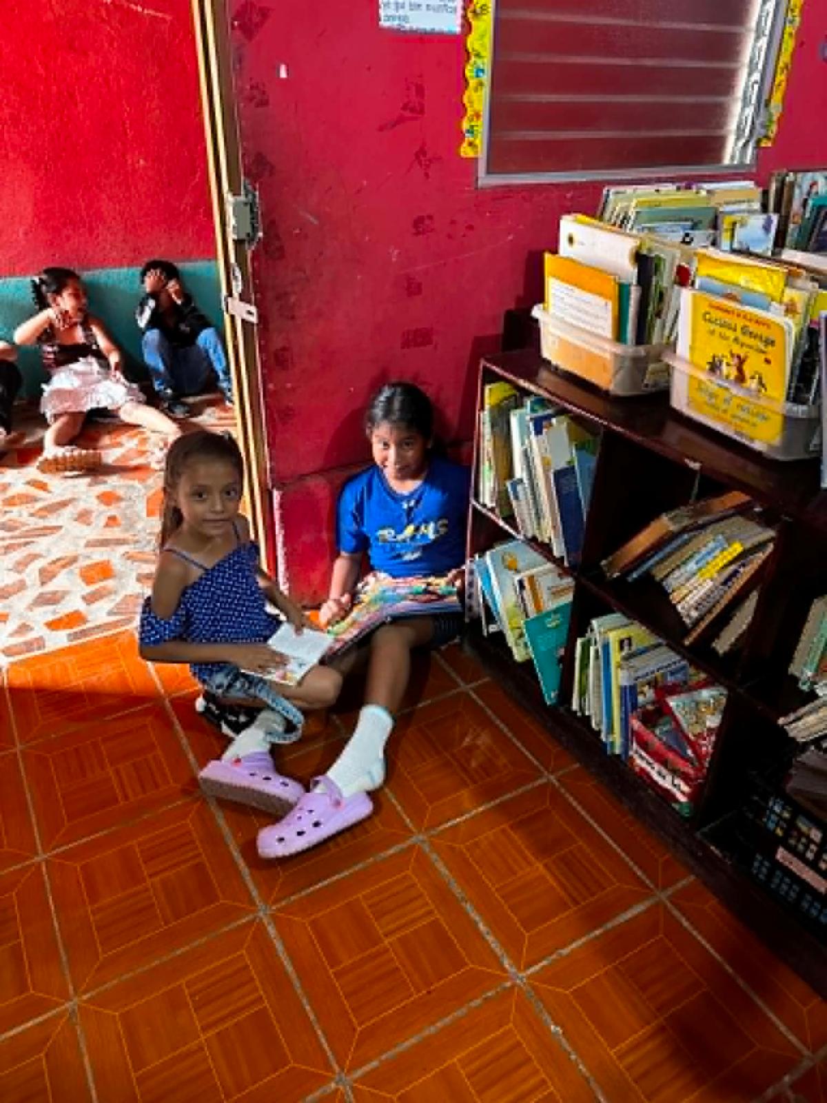 Candid3 - Neighbors to Nicaragua Two girls sitting on the floor reading books in a small library corner, with bright pink walls and a shelf of magazines nearby nearby readers and a patterned orange floor tile.
