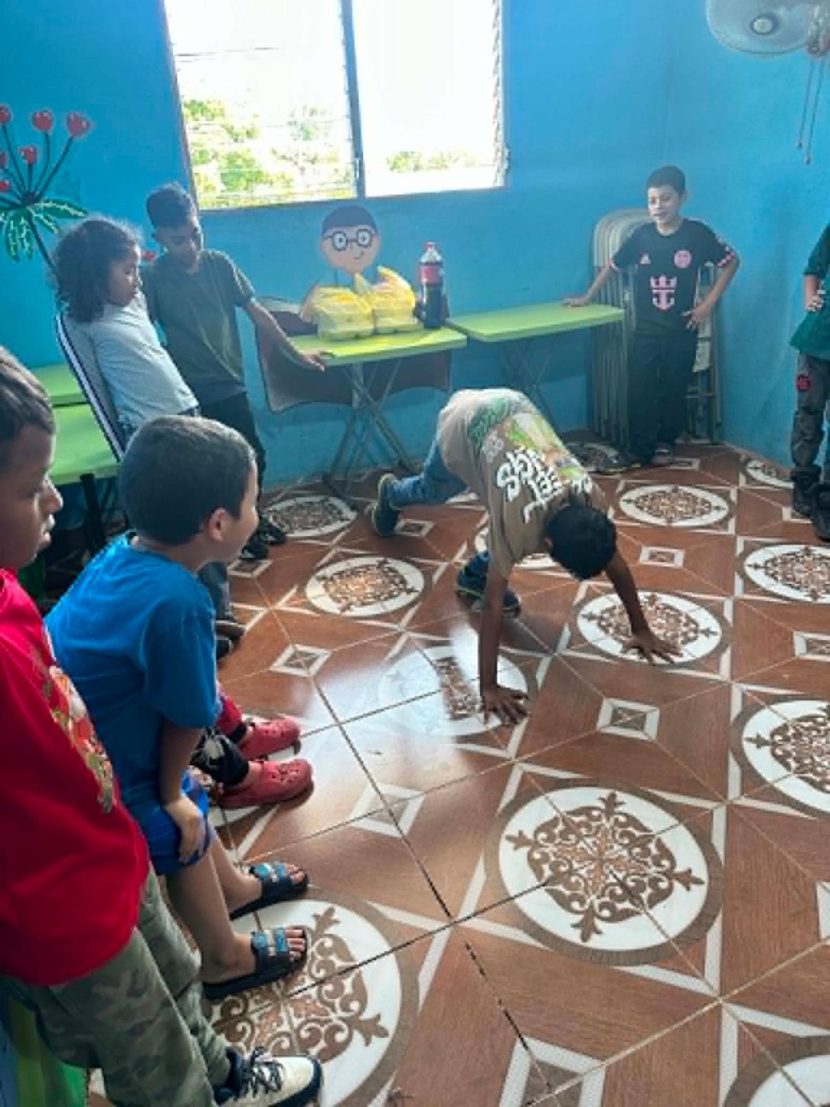 Candid4 - Neighbors to Nicaragua Group of children in a blue classroom watching a boy perform push-ups on a patterned tile floor.