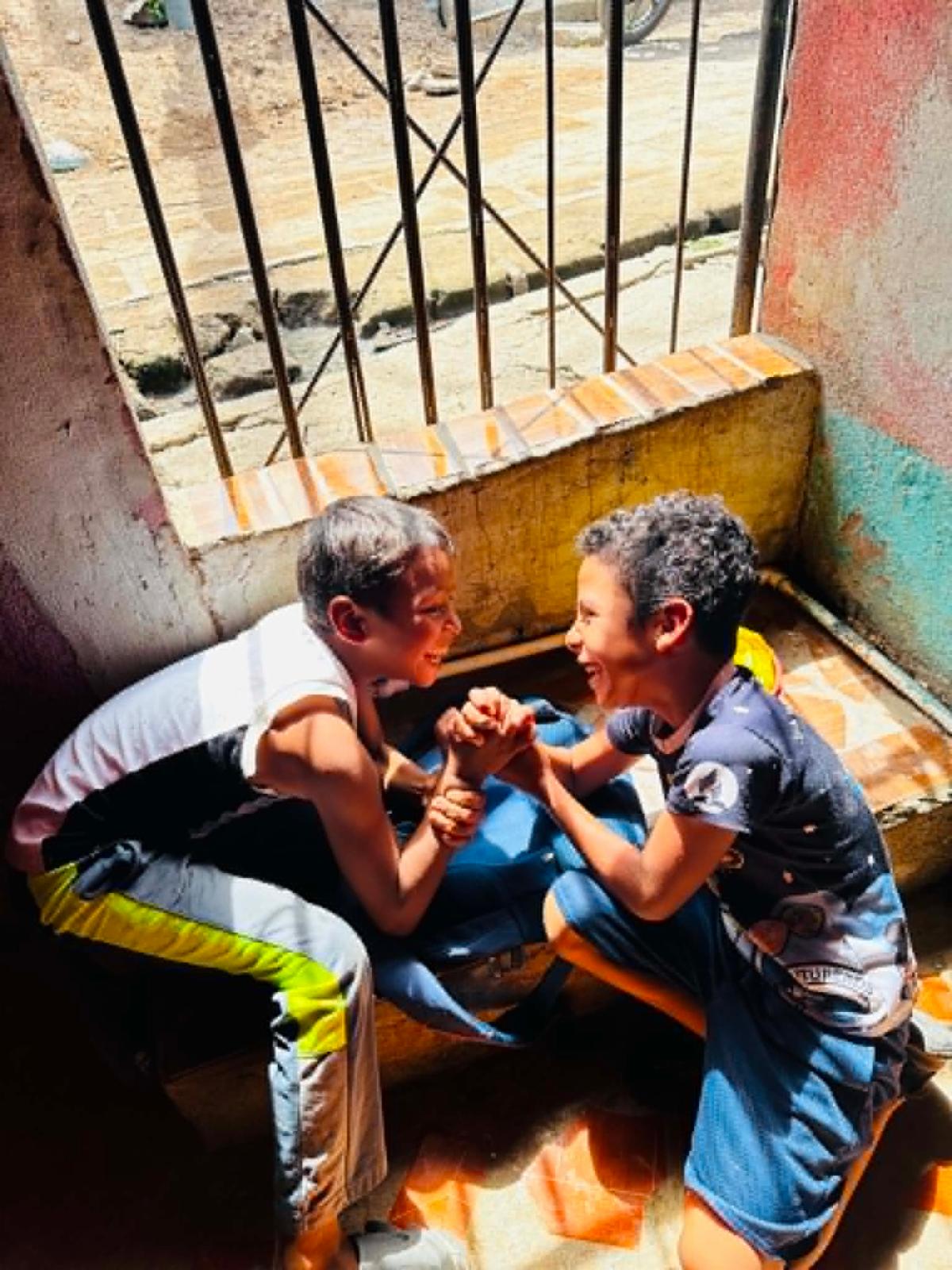 Candid5 - Neighbors to Nicaragua Two boys arm-wrestling and smiling in a sunny, enclosed space with colorful walls and a metal railing behind them.