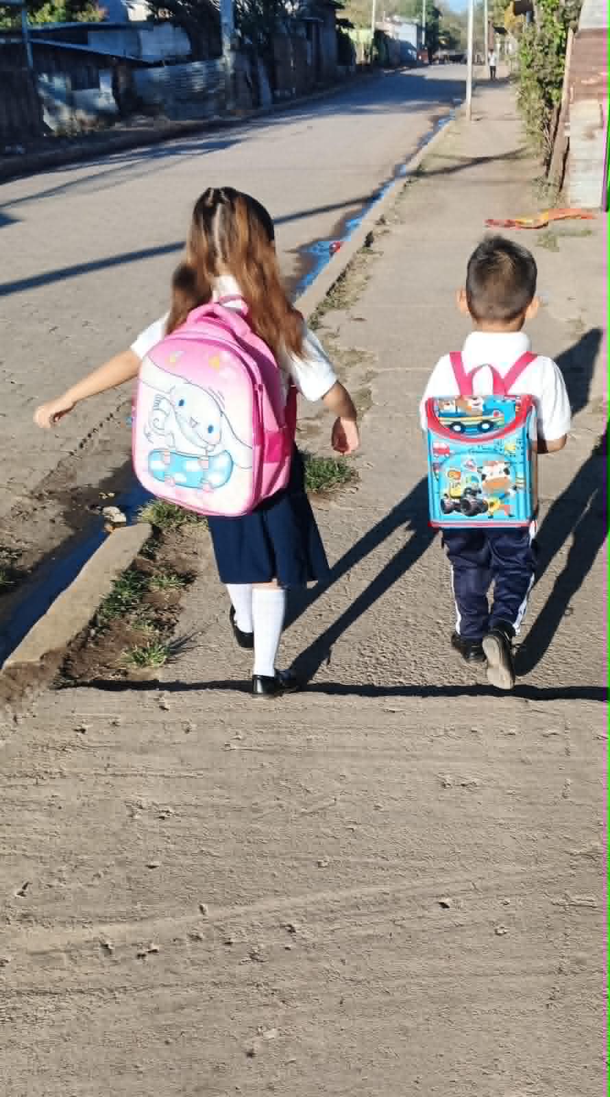 Candid6 - Neighbors to Nicaragua Two young children walk away from the camera on a sunny sidewalk, each wearing a colorful backpack (pink with a cartoon character, blue with trucks).