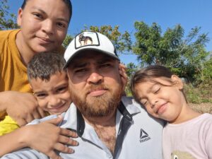 Garcia Family - Neighbors to Nicaragua Family selfie outdoors: bearded man in a white cap surrounded by two children and a woman on a sunny day with trees in the background.