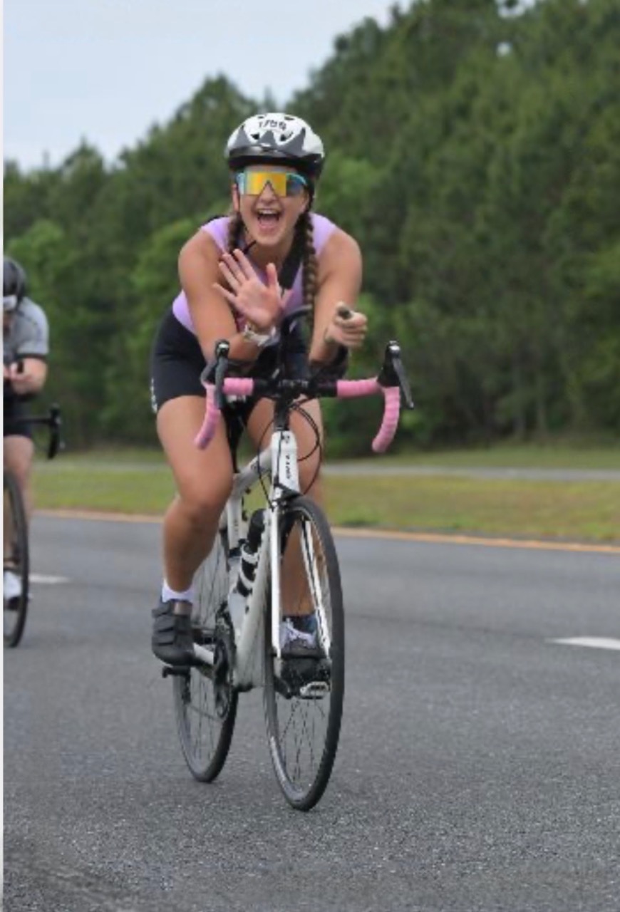 IronMan1 - Neighbors to Nicaragua Cyclist in a purple top and helmet rides a road bike, waving and smiling during a race with pink handlebars.
