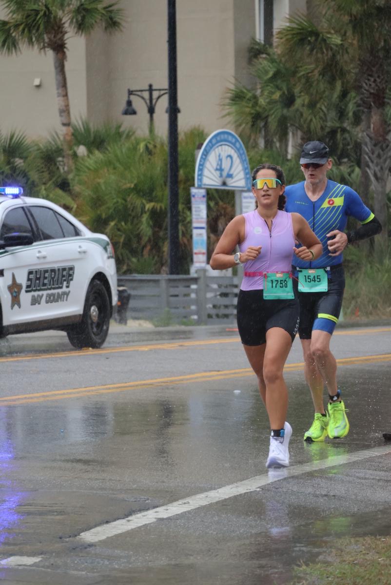 IronMan2 - Neighbors to Nicaragua Two runners race in the rain on a wet road, bib numbers 1758 and 1545, with a sheriff’s SUV parked nearby.