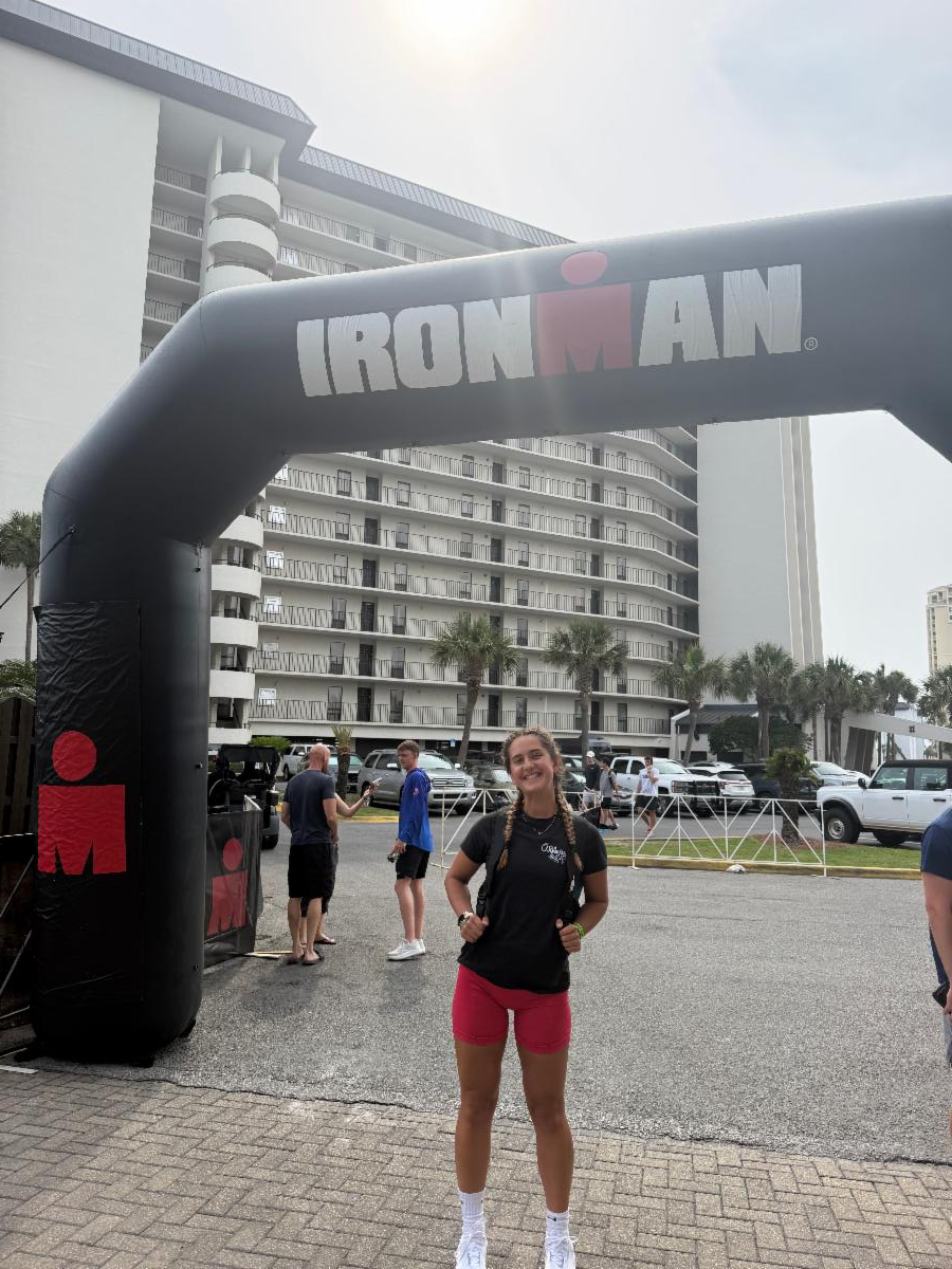 IronMan3 - Neighbors to Nicaragua Young woman in a black T-shirt and pink shorts posing under an Ironman inflatable arch at an outdoor race event by a hotel.