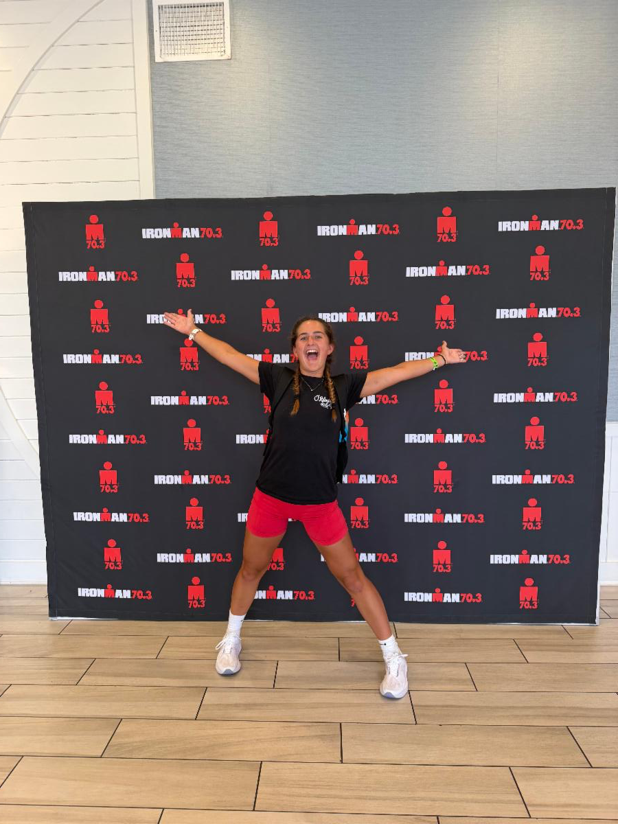 IronMan4 - Neighbors to Nicaragua Woman with two braids, white sneakers, and black shirt posing energetically in front of an Ironman 70.3 backdrop with arms spread wide in a victory pose.