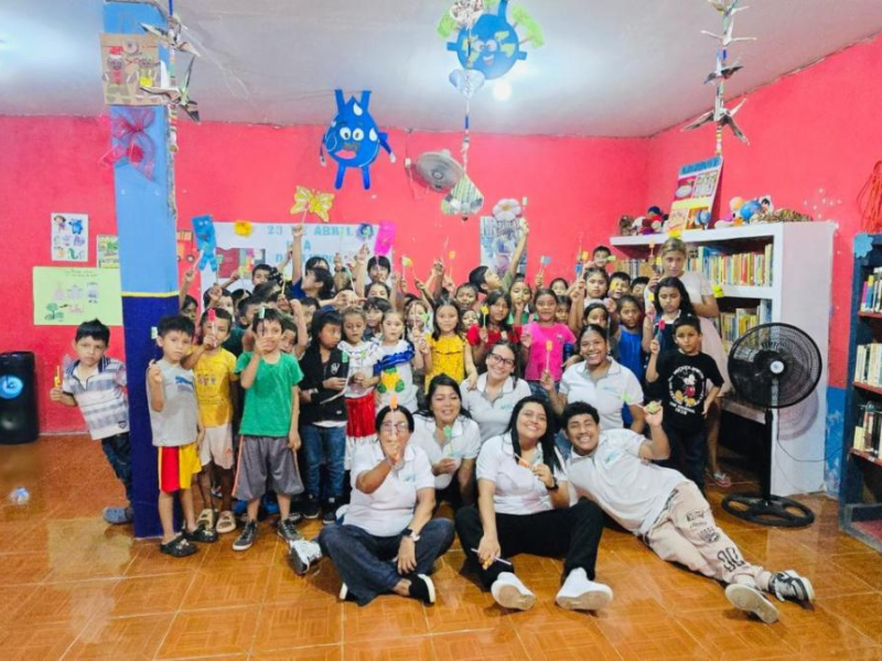 Group of children and adults posing for a group photo in a colorful classroom/library, with pink walls, shelves, and hanging decorations.
