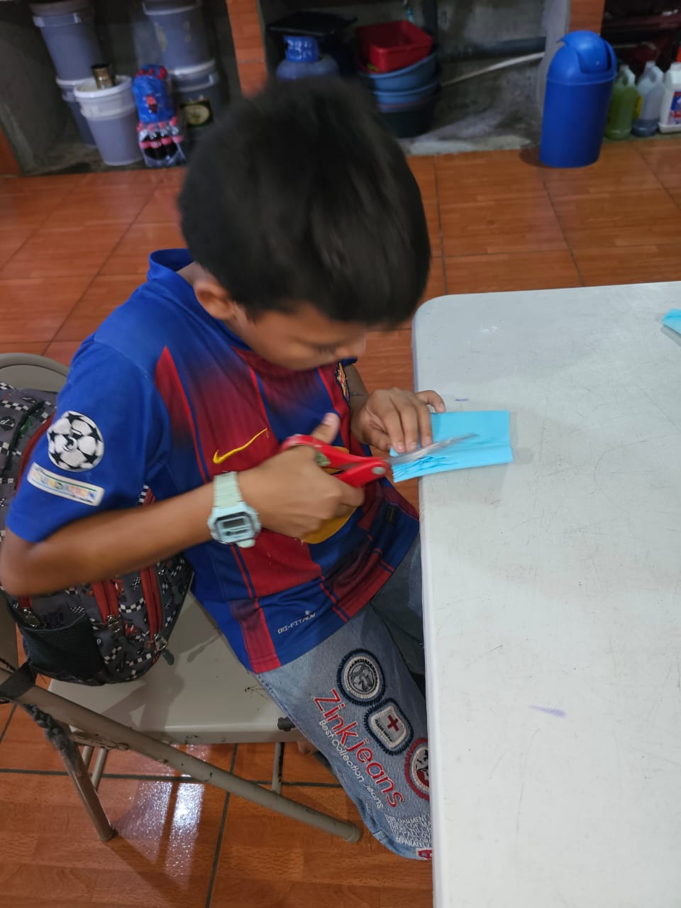 MothersDay1 - Neighbors to Nicaragua Young boy in a blue and red shirt cuts blue paper with scissors at a table during a craft activity.