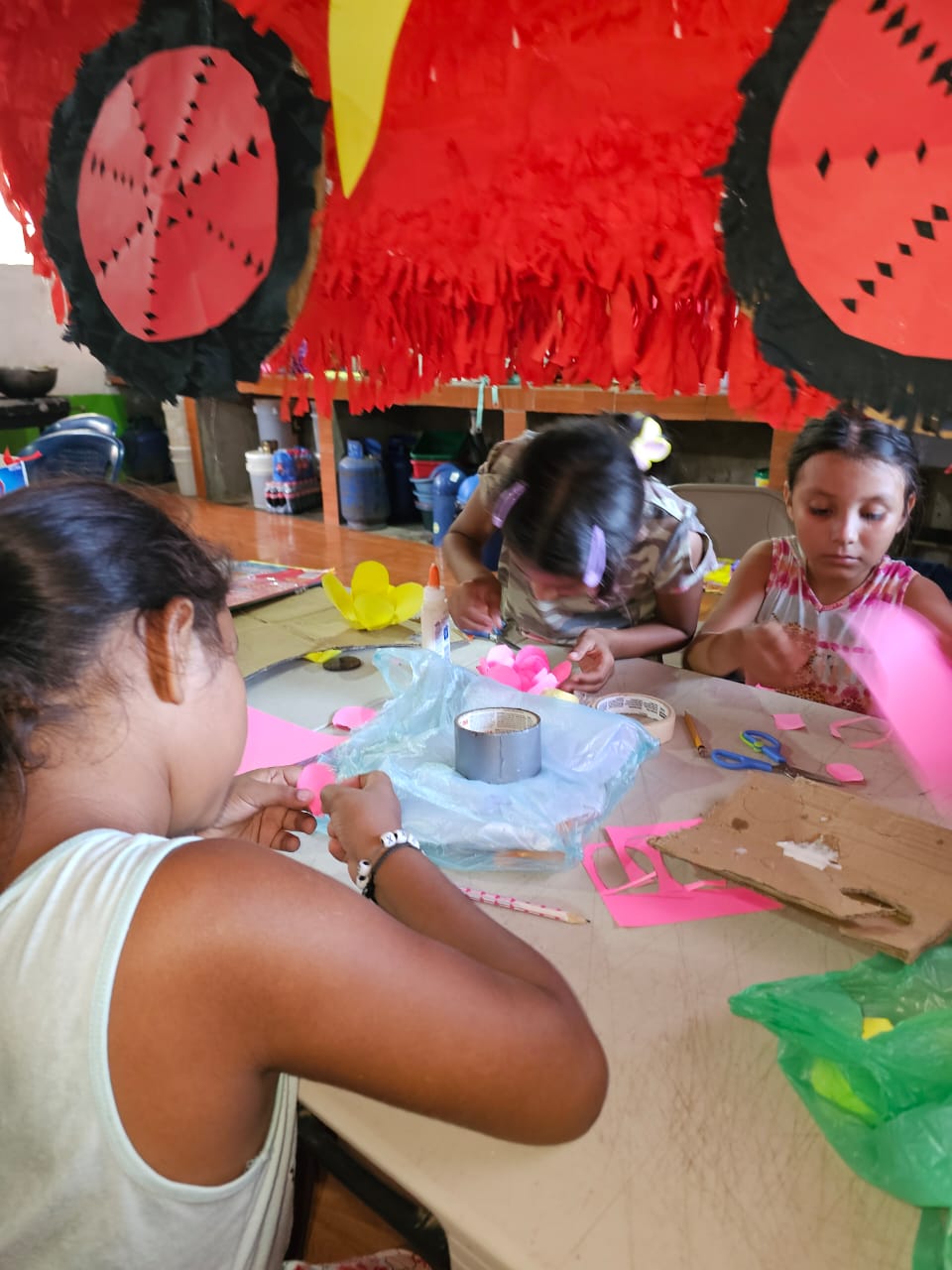 MothersDay2 - Neighbors to Nicaragua Children sitting around a table in a bright craft room, cutting and gluing pink paper for a DIY project as red decorations hang overhead.
