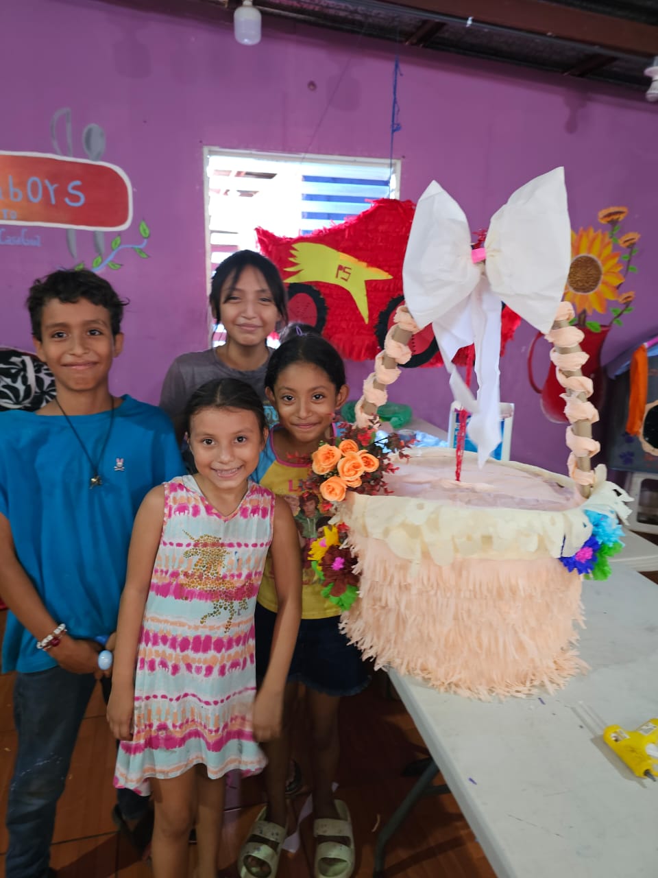 MothersDay3 - Neighbors to Nicaragua Four children stand together in a purple room next to a large decorative basket adorned with pink fringe, orange flowers, and a big white bow on a table.