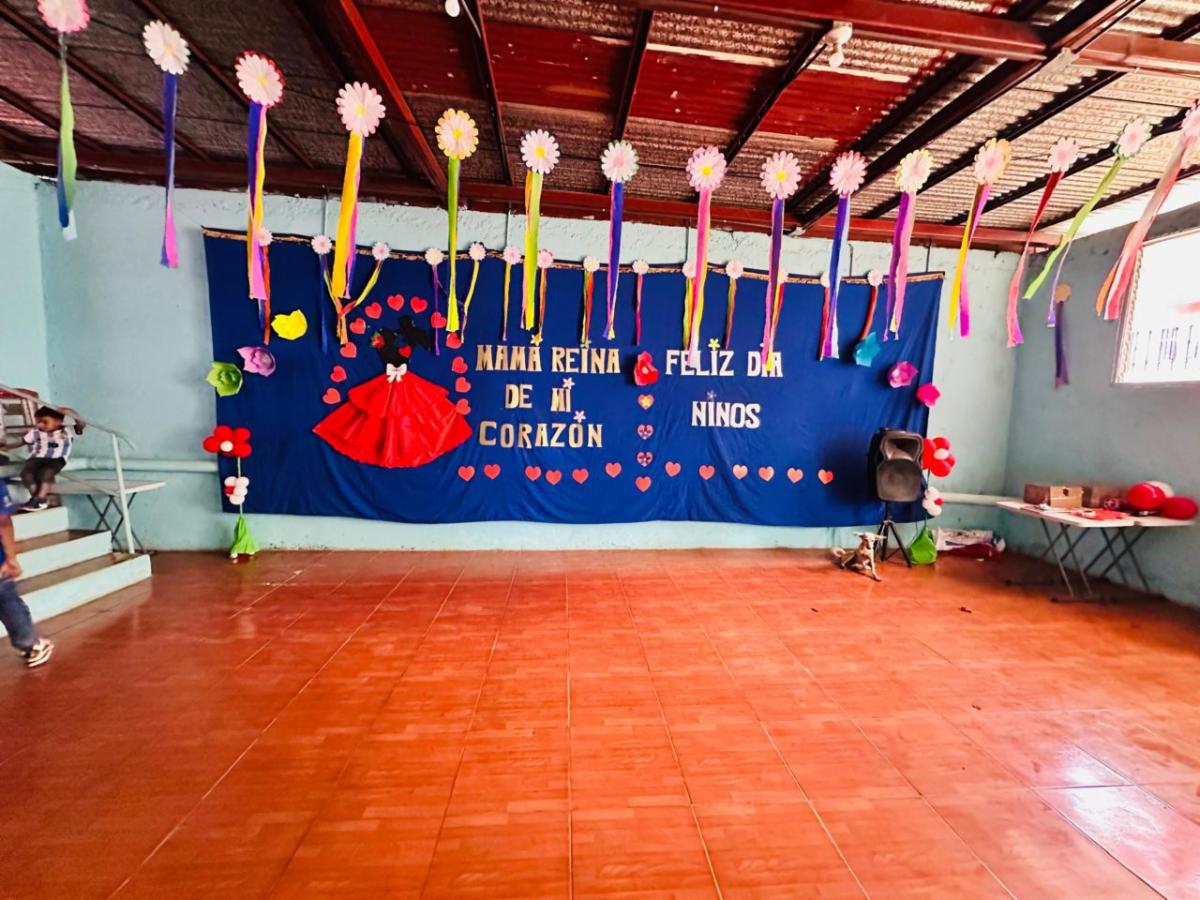 MothersDay4 - Neighbors to Nicaragua Festively decorated indoor space for a Children’s Day event, with a blue banner reading Feliz Día de Niños, colorful streamers, and paper flowers-inspired decorations.",