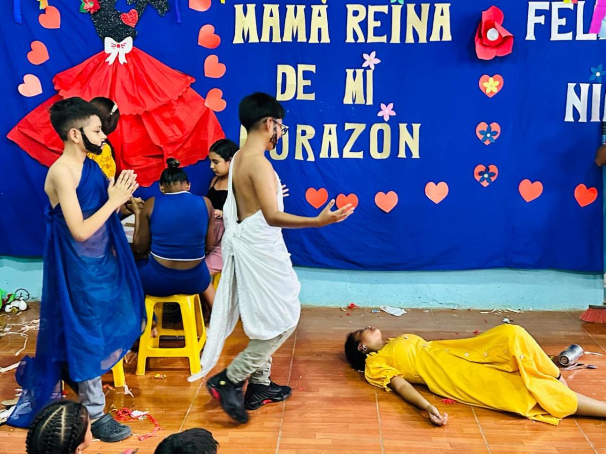 MothersDay5 - Neighbors to Nicaragua Group of children in a classroom stage, some in robes, one child in yellow dress lying on the floor while others walk by, against a blue backdrop with hearts and a banner.