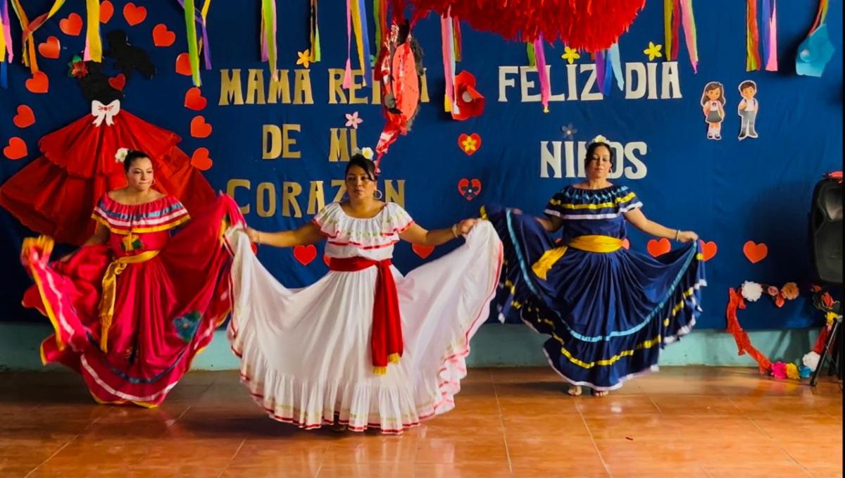 MothersDay6 - Neighbors to Nicaragua Three women in colorful traditional dresses perform a folkloric dance on a decorated stage for Children's Day, with a blue backdrop and paper hearts behind them.