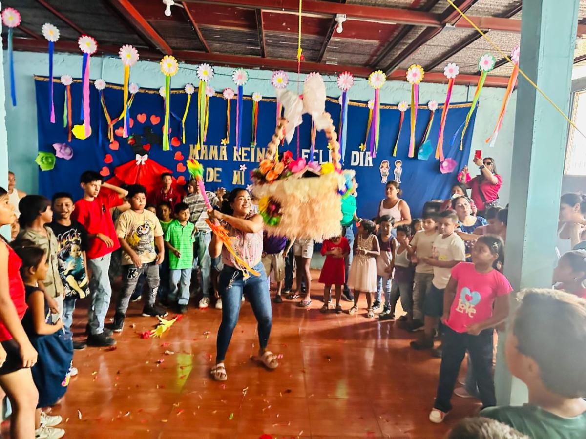 MothersDay8 - Neighbors to Nicaragua Children form a circle around a woman swinging a colorful piñata in a decorated party hall with paper flowers and ribbons.