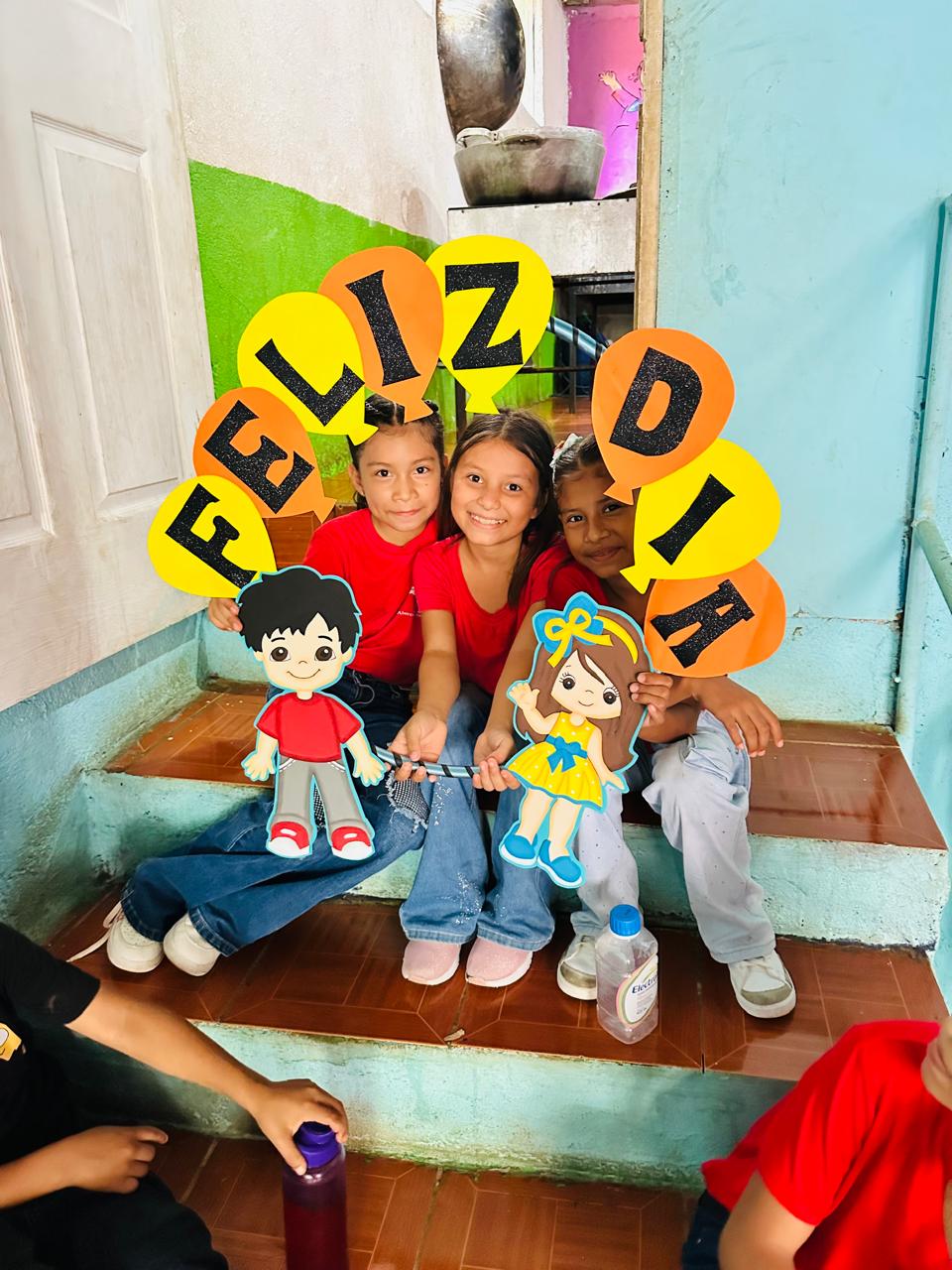 MothersDay9 - Neighbors to Nicaragua Group of smiling children sitting on steps, holding a colorful cutout banner that spells 'FELIZ DIA' with cartoon boy and girl figures.