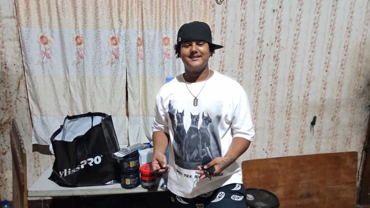 Ramon1 - Neighbors to Nicaragua Man wearing a white graphic T-shirt and black cap, smiling in a cluttered room, holding small tools near a table with a black tote bag.
