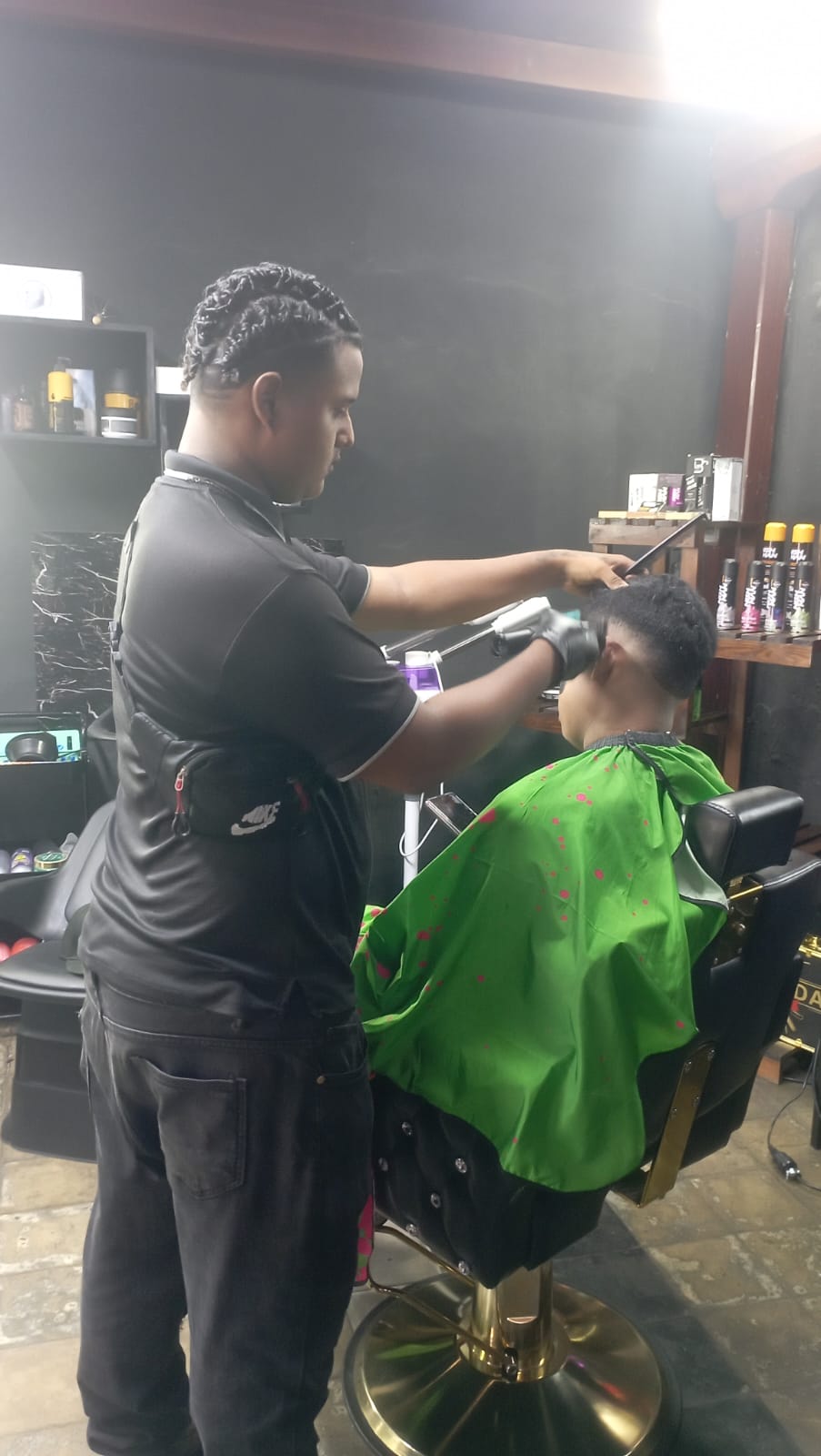 Ramon3 - Neighbors to Nicaragua Barber in a black shirt trims a client's hair with clippers while the client wears a green cape in a barber chair.