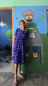 Girl in a blue floral dress stands beside a large mural of a student at a desk on a green and blue classroom wall.
