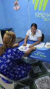 Woman in a white polo helps a girl with worksheets at a blue desk in a bright blue room.