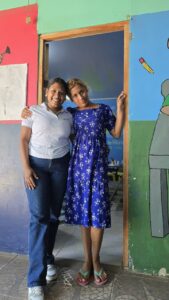 Safe Places 3 - Neighbors to Nicaragua Two women stand in a doorway inside a colorful mural room, smiling; one wears a white polo and jeans, the other a blue floral dress.