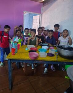 Sponsorship May - Neighbors to Nicaragua Group of smiling children standing behind a yellow table filled with bowls, cups, and a large pot in a colorful cooking classroom/space.”