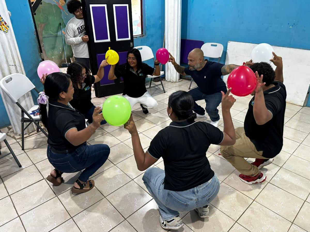 Teachers 2 - Neighbors to Nicaragua Group of adults in a circle, squatting and holding colorful balloons during a team-building activity in a blue-walled room.