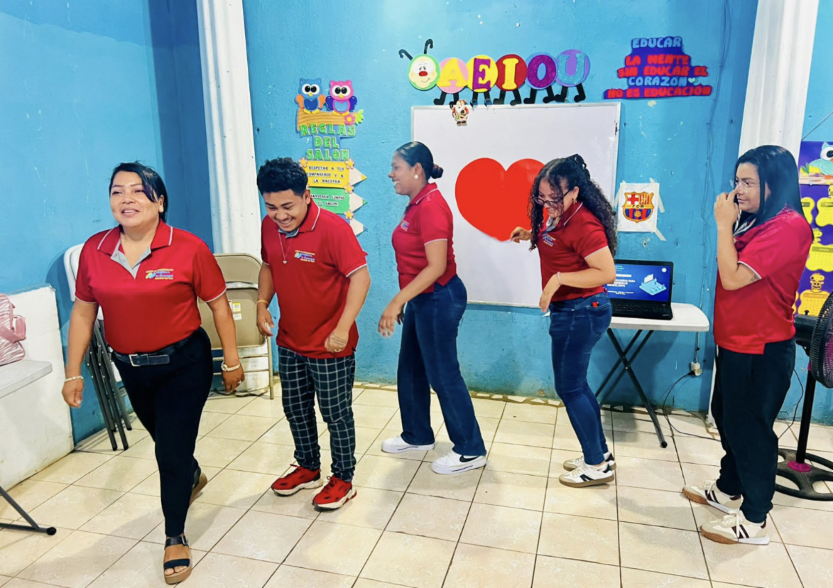 Teachers 3 - Neighbors to Nicaragua Five people in red polo shirts walk in a line in a blue-walled classroom with posters and a large red heart on the wall behind them.