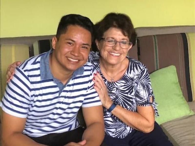 Young man in a blue-and-white striped polo sits beside an elderly woman with glasses; both smile on a striped couch in a lime-w colored room.