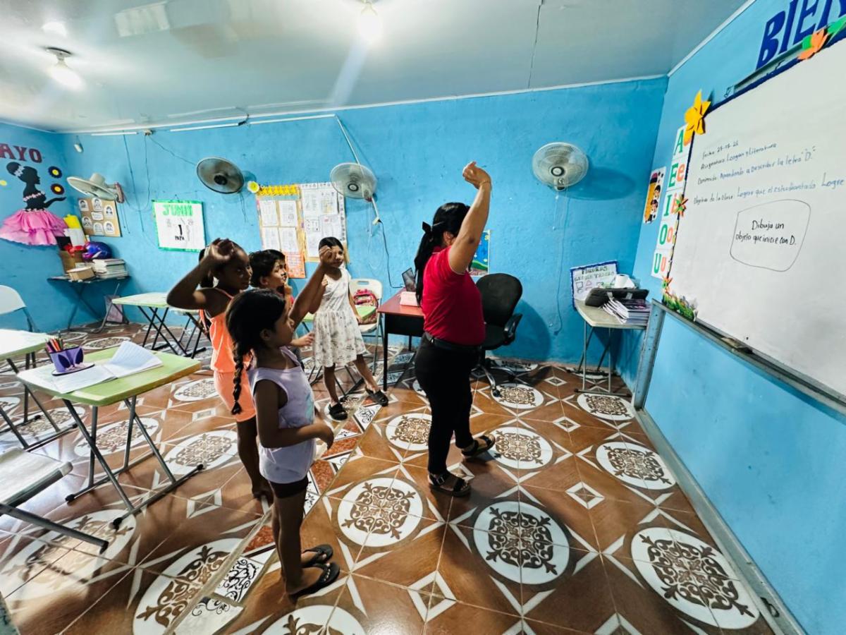 Teacher in a red shirt leads a group of children in an arm-raising activity in a bright blue classroom with tiled floor and desks nearby