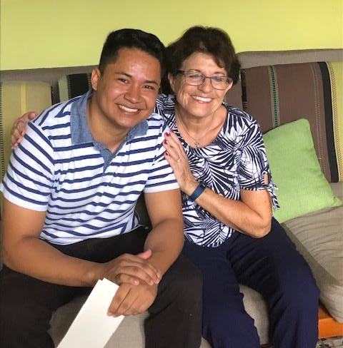 Smiling man and an older woman sit close together on a couch, arms around each other, posing for a photo in a living room.
