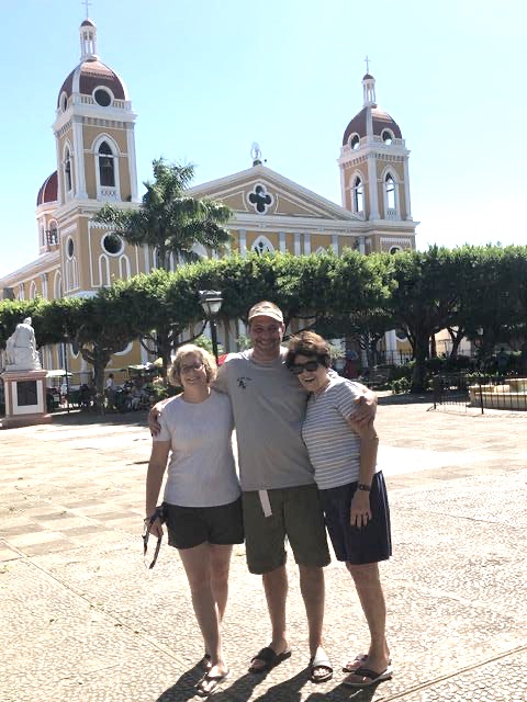 Three adults stand arm in arm in a sunny plaza, with a yellow church featuring twin towers in the background.