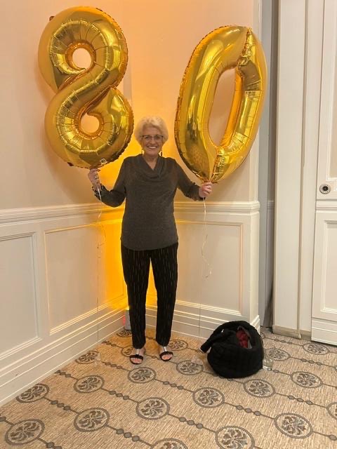 Person standing in a neutral hallway, holding large gold balloons shaped like 8 and 0 to celebrate an 80th birthday.
