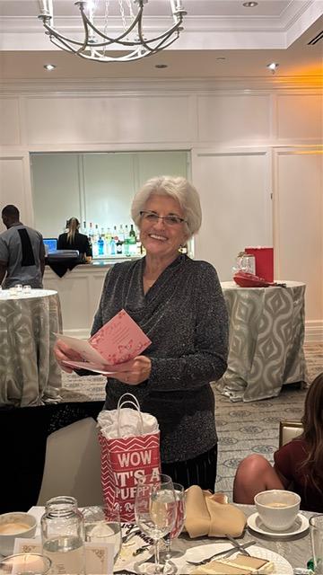 Smiling older woman with white hair and glasses holds pink cards at a formal reception, standing beside a staged gift table in a banquet hall.