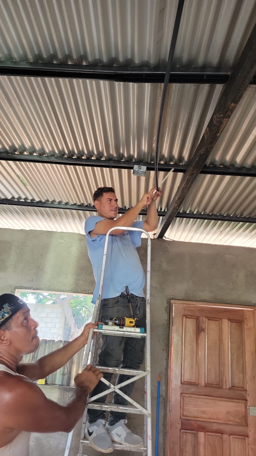 Two men install electrical wiring on a corrugated metal ceiling; one stands on a ladder guiding a cable while another holds the ladder steady with a drill nearby.