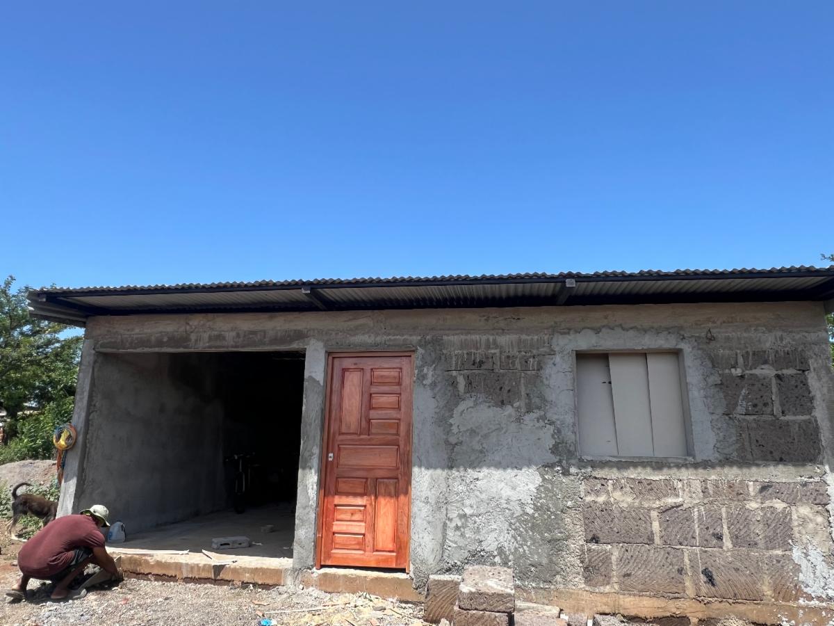 Worker kneeling near unfinished concrete house with a red wooden door and a window under a bright blue sky; a dog nearby on the left.