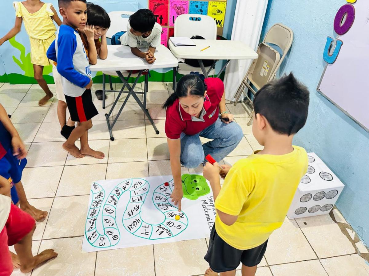 Teacher kneels on the tiled floor guiding a group of children as they play a large snake-and-ladder style math game laid out on a white poster.