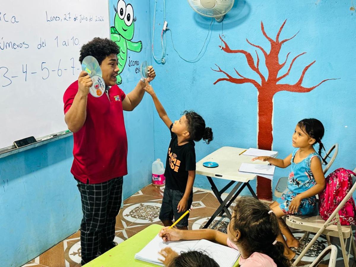 Teacher in a red shirt holds two CDs up toward a young student in a bright classroom while other kids write at desks in front of a blue wall with a tree mural and a whiteboard.