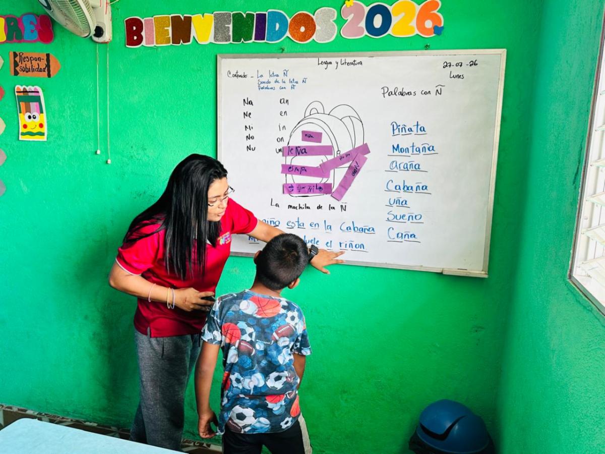 Teacher helps a student at a green classroom wall, pointing to a whiteboard with words and a purple diagram.