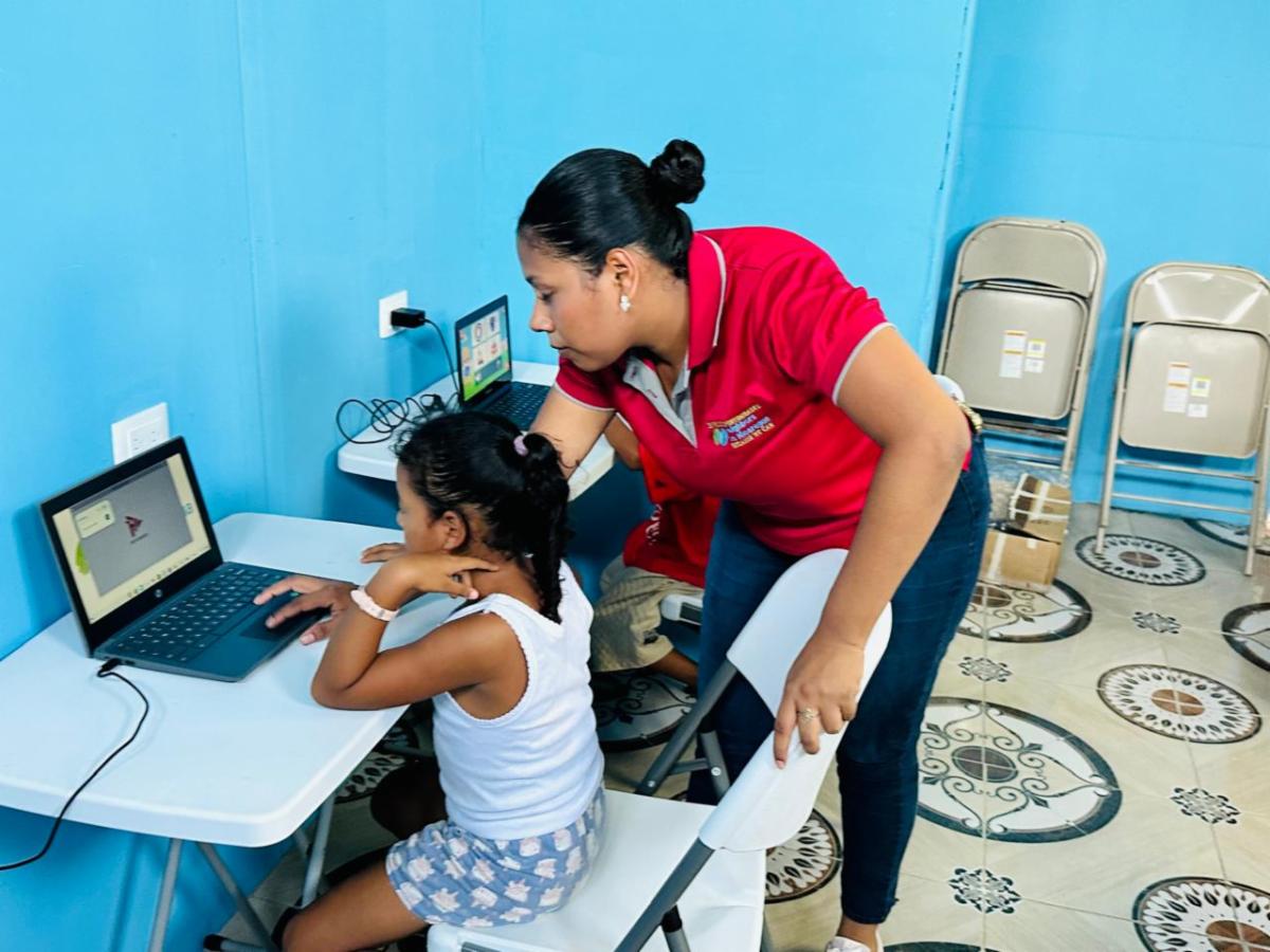 Woman in a red shirt assists a young girl as she works on a laptop at a blue-walled computer station.