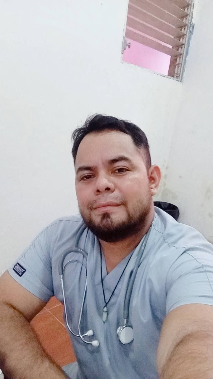 Male healthcare worker in light blue scrubs with stethoscope, taking a selfie in a bright room.