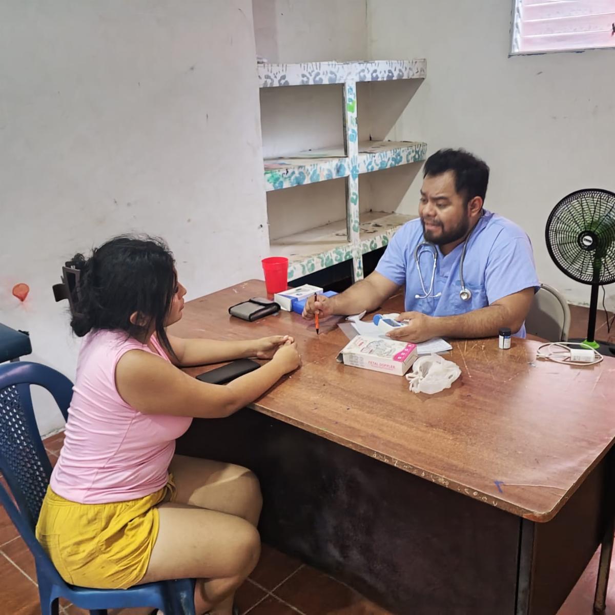 Male doctor in blue scrubs with a stethoscope consults a female patient at a wooden desk in a clinic.