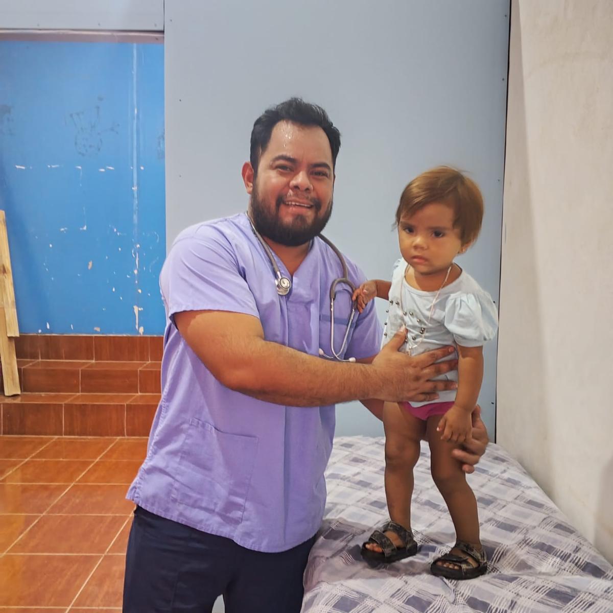 Smiling male medical professional in lavender scrubs holds a young girl on an examination table (clinic setting).