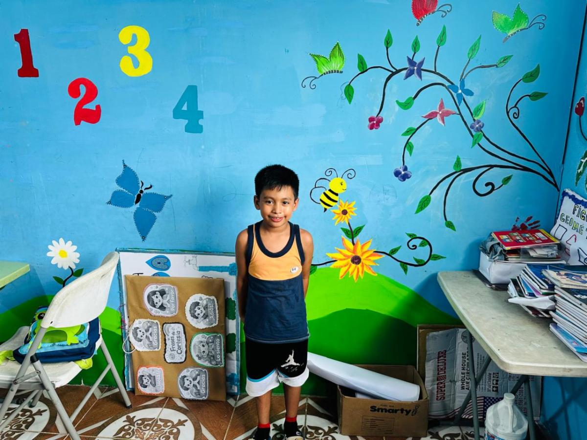 Smiling boy in a sleeveless shirt stands in a bright muraled classroom corner with numbers on the blue wall, a painted tree, flowers, and butterflies.