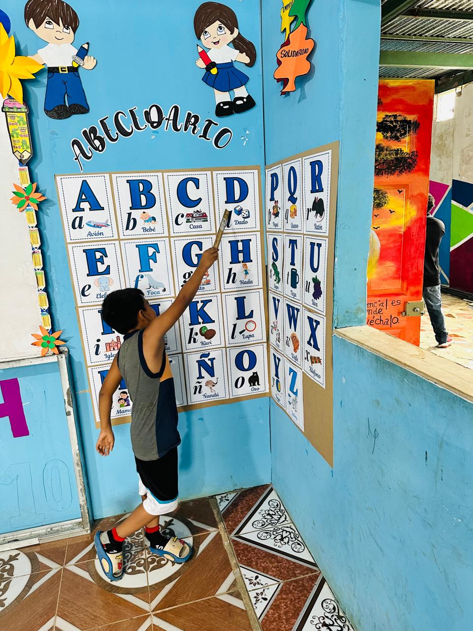 Boy wearing a sleeveless shirt points at a grid of large alphabet cards (A–Z) on a blue classroom wall, with colorful pictures beside each letter.